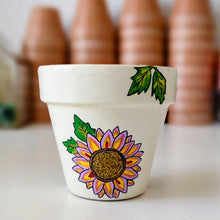 Load image into Gallery viewer, A small white painted flower pot with a colorful sunflower design and green leaves, set on a white surface with blurred terracotta pots in the background.