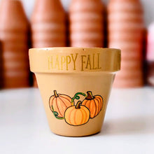 Load image into Gallery viewer, A small hand-painted terracotta pot with 'HAPPY FALL' and pumpkins on it, with other pots blurred in the background.

